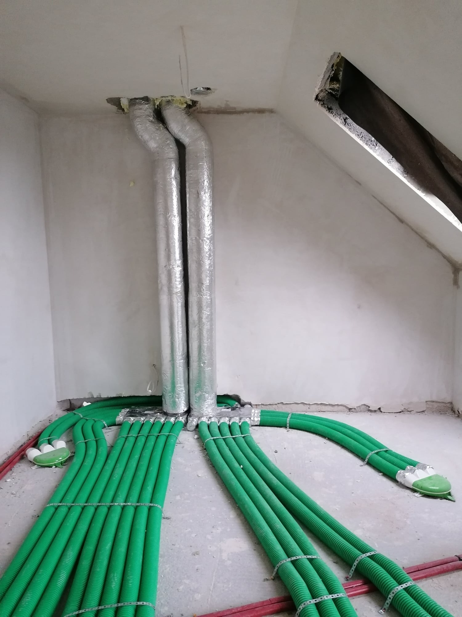 Zielone rury wentylacyjne ułożone na podłodze w surowym wnętrzu poddasza, połączone z dwoma srebrnymi, izolowanymi przewodami wychodzącymi z sufitu.