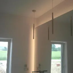 Nowoczesna łazienka z trzema pionowymi lampami wiszącymi w kształcie tub, lustro na całej ścianie z podświetleniem LED, widok na zielone pole przez okno.