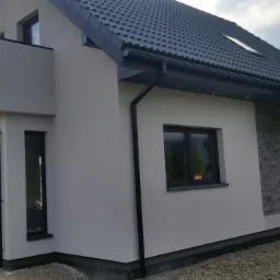 Nowoczesny dom jednorodzinny z szarą elewacją, ciemną dachówką i grafitowymi rynnami, widok z zewnątrz na ukośną ścianę z wykończeniem imitującym kamień.