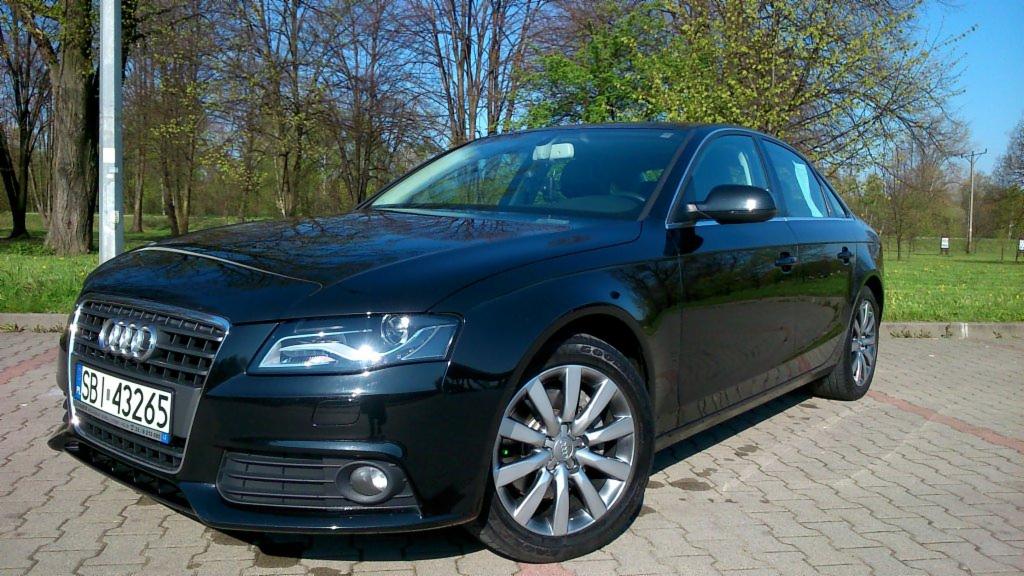 Czarny samochód osobowy Audi A4 na parkingu z kostki brukowej, widoczny numer rejestracyjny SBI 43265, w tle drzewa i trawnik.