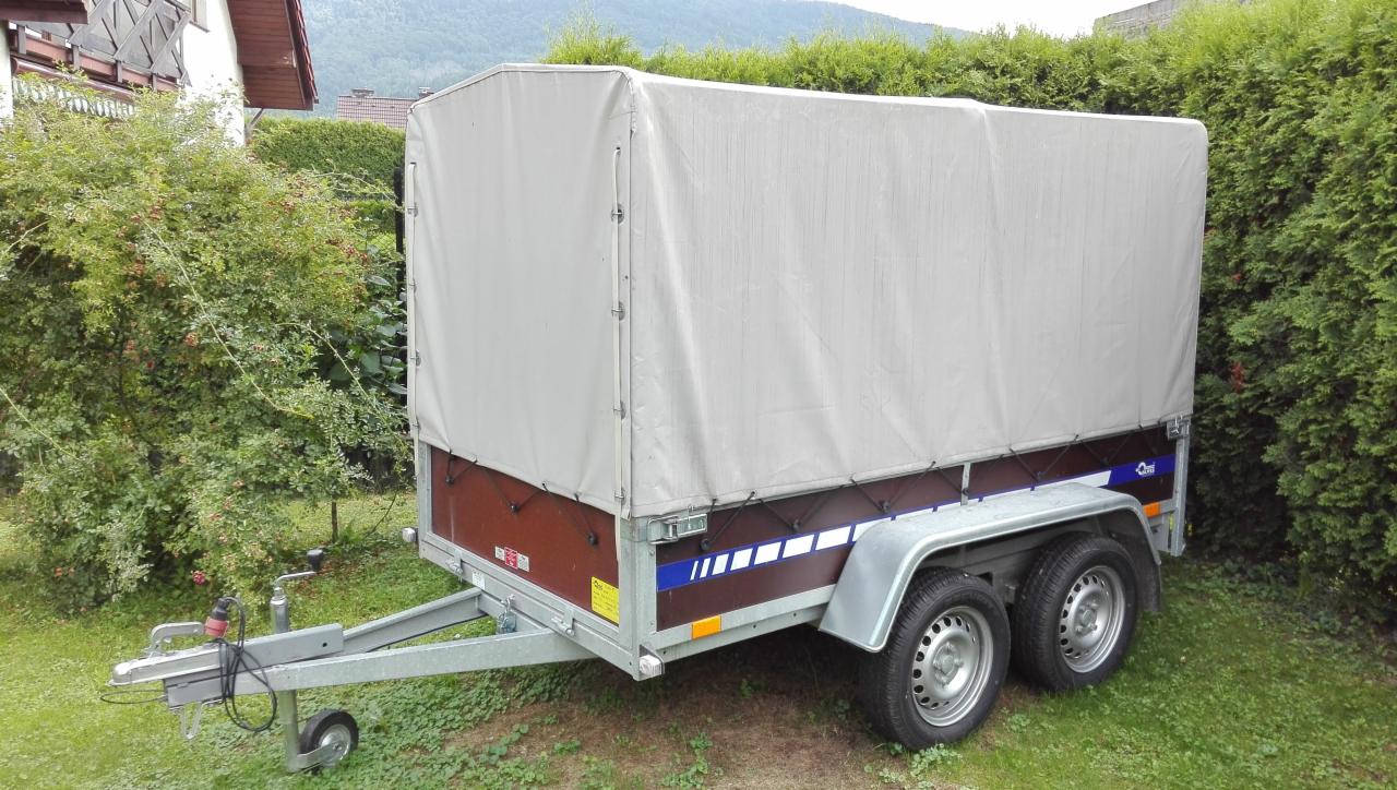 Przyczepa dwuosiowa z plandeką w kolorze szarym, zaparkowana na trawniku przed zielonymi krzewami i budynkiem. Widoczne opony, błotniki, dyszel z zaczepem i podpórką. Plandeka czysta, bez napisów.