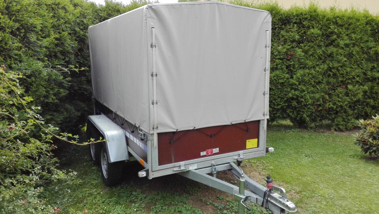 Przyczepa z plandeką w kolorze szarości, zaparkowana na trawie przed żywopłotem.