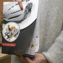 Osoba trzyma w dłoniach otwarty katalog z grafiką samochodu i części samochodowych, prezentujący ofertę handlową. Widoczne strony z logo firm i tekstem.