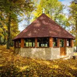 Ośmioboczna altana z brązową dachówką bitumiczną, kamienną podstawą i drewnianą stolarką okienną, otoczona jesiennym lasem z opadłymi liśćmi.