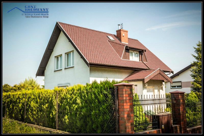 Dom jednorodzinny z brązowym dachem, widoczny zza żywopłotu i ogrodzenia. Dachówka ceramiczna, komin z cegły, okno dachowe. Jasne niebo w tle.