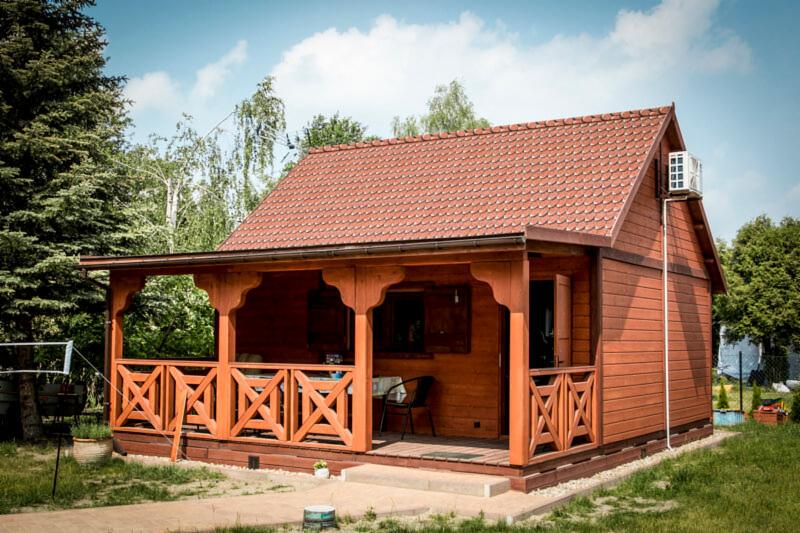Drewniany domek letniskowy z czerwonym dachem z dachówki karpiówki i drewnianą werandą, widok z zewnątrz w słoneczny dzień.