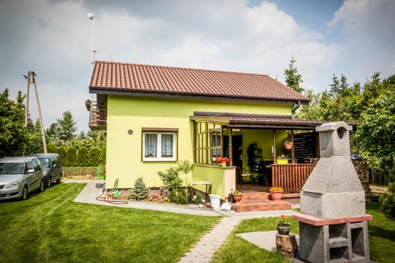 Dom jednorodzinny z brązową dachówką, zieloną elewacją i zadaszonym tarasem. Na pierwszym planie murowany grill, a w tle zaparkowane samochody.