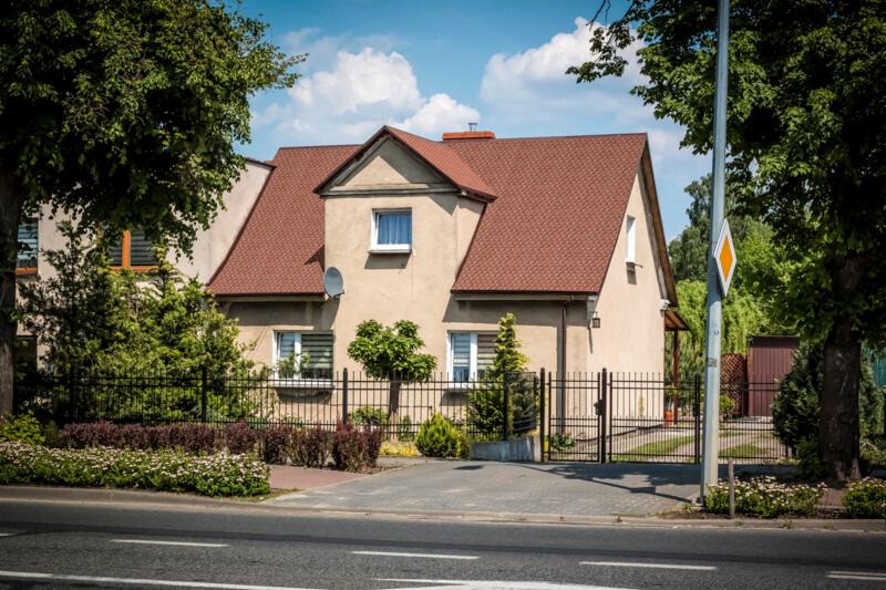 Dom jednorodzinny z brązowym dachem z dachówki, ogrodzony czarnym płotem, widok z ulicy w słoneczny dzień.