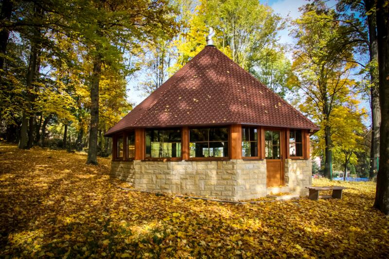 Ośmioboczna altana z brązową dachówką bitumiczną, kamienną podstawą i drewnianą stolarką okienną, otoczona jesiennym lasem z opadłymi liśćmi.