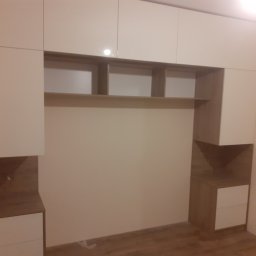 MEBLEART - Zabudowa meblowa na wymiar w pokoju, z górnymi szafkami w kolorze kremowym i elementami w odcieniach drewna, z otwartymi półkami nad miejscem na łóżko, widoczne niedoskonałości oświetlenia.