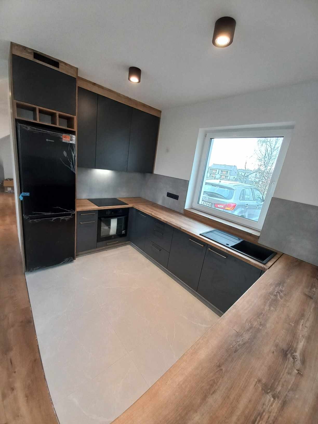 Nowoczesna, szara kuchnia z drewnianymi blatami, czarnym zlewem i sprzętem AGD, oświetlona punktowymi lampami sufitowymi, z widokiem na samochód za oknem.