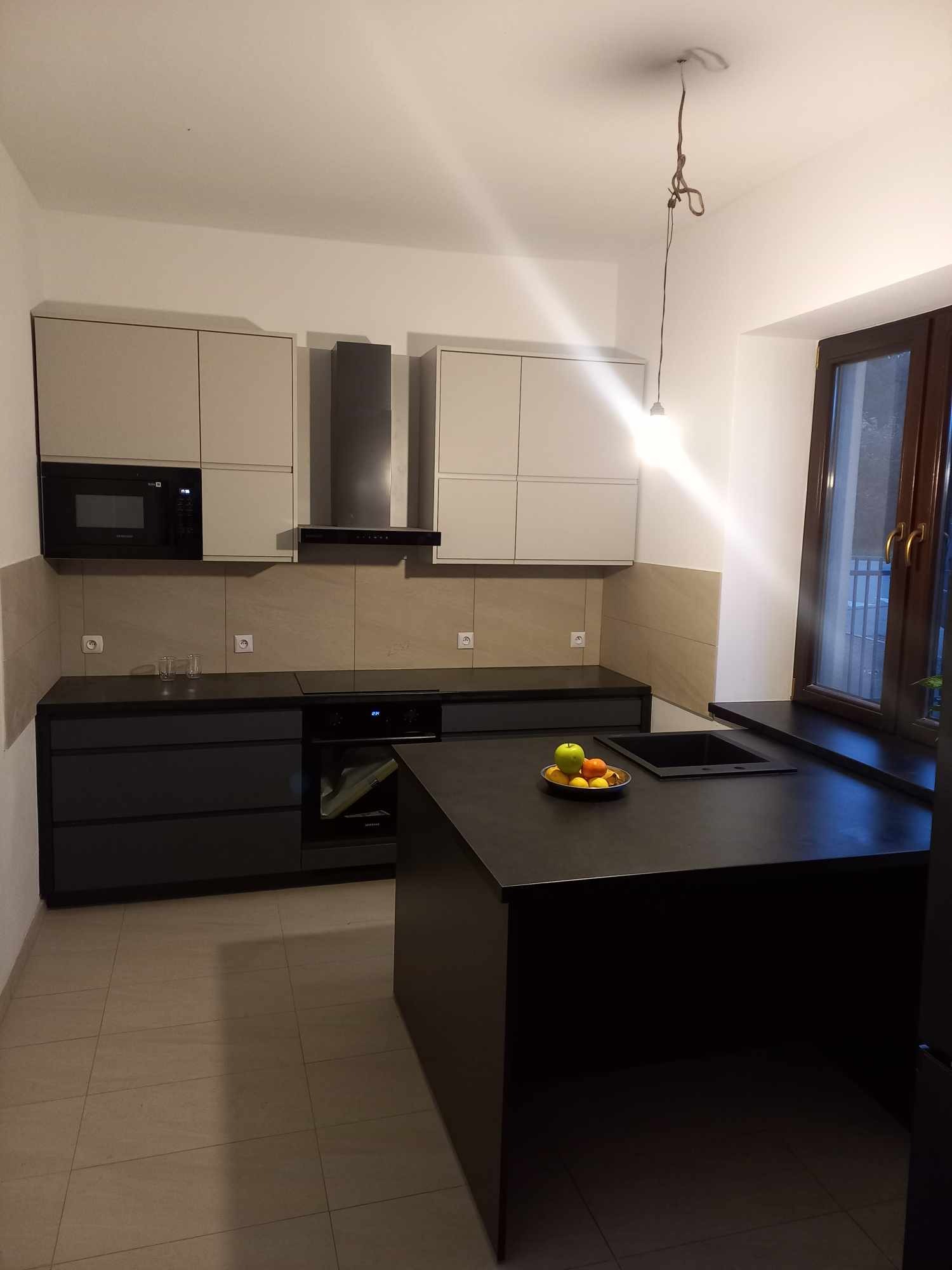 Nowoczesna kuchnia z wyspą i czarnymi blatami, widoczny owoc w misce na wyspie, niedokończona instalacja lampy sufitowej.