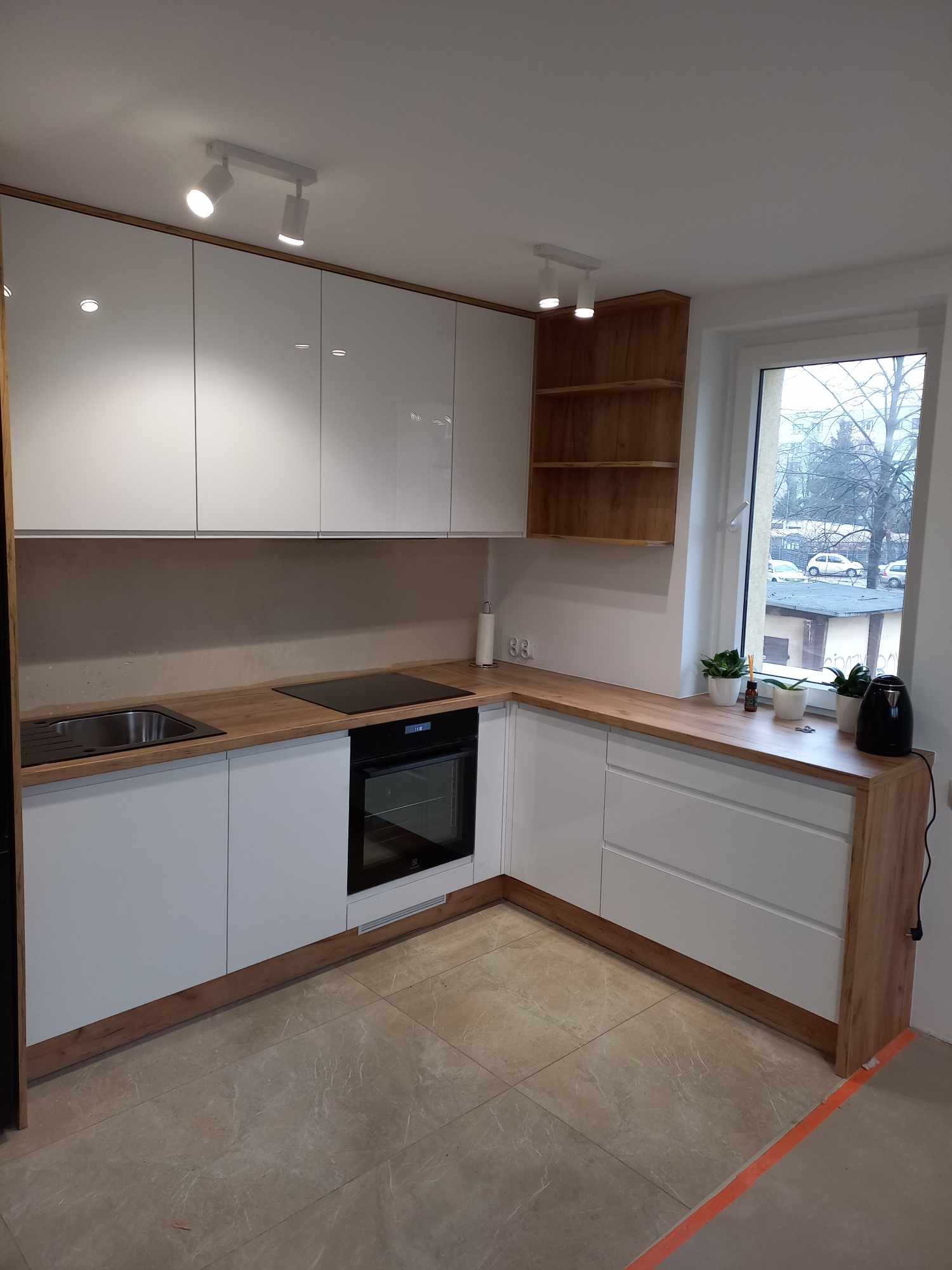 Nowoczesna, narożna kuchnia z białymi, połyskującymi frontami szafek i drewnianym blatem, oświetlona reflektorami; widok na okno z roślinami doniczkowymi.