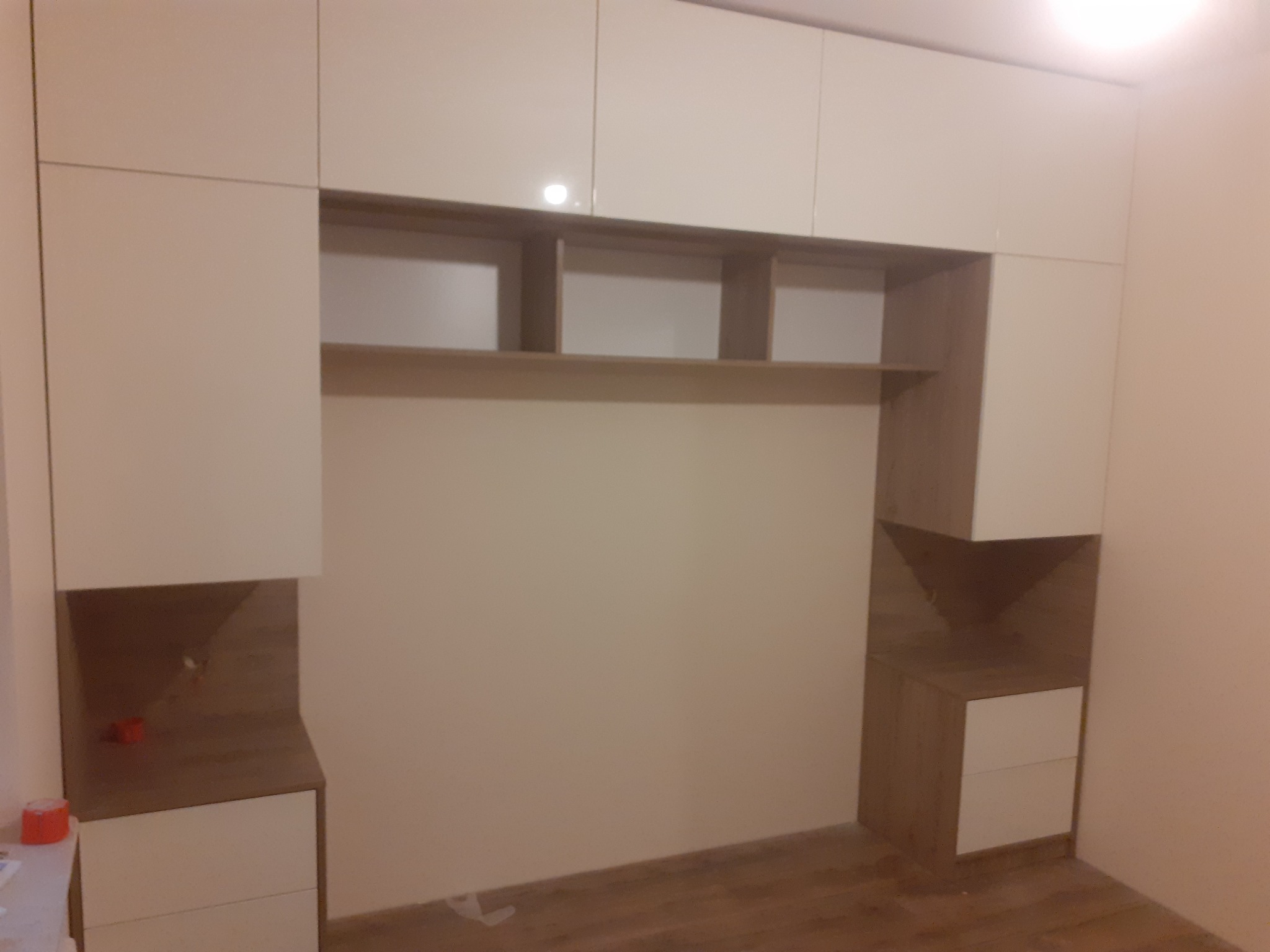 Zabudowa meblowa na wymiar w pokoju, z górnymi szafkami w kolorze kremowym i elementami w odcieniach drewna, z otwartymi półkami nad miejscem na łóżko, widoczne niedoskonałości oświetlenia.