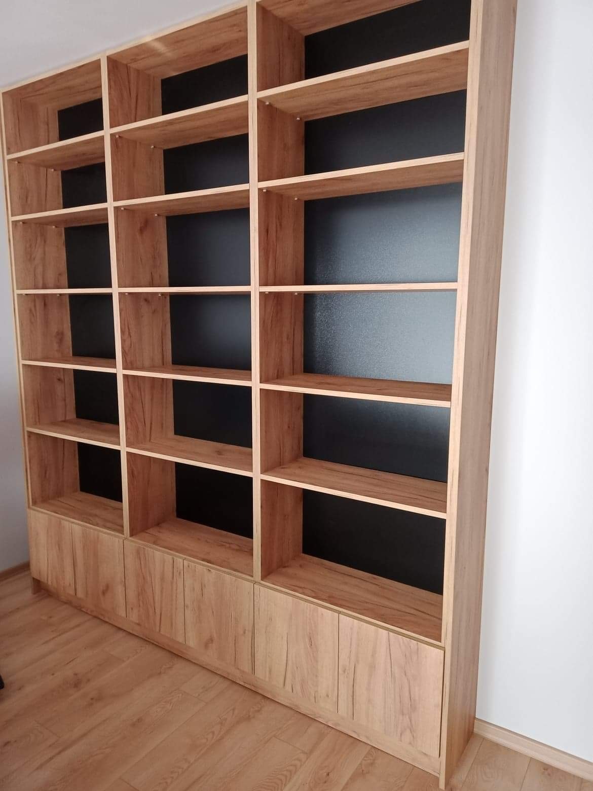 Duża, drewniana biblioteczka z półkami i szafkami na dole, z czarnym tłem wewnątrz półek, ustawiona na drewnianej podłodze przy białej ścianie.