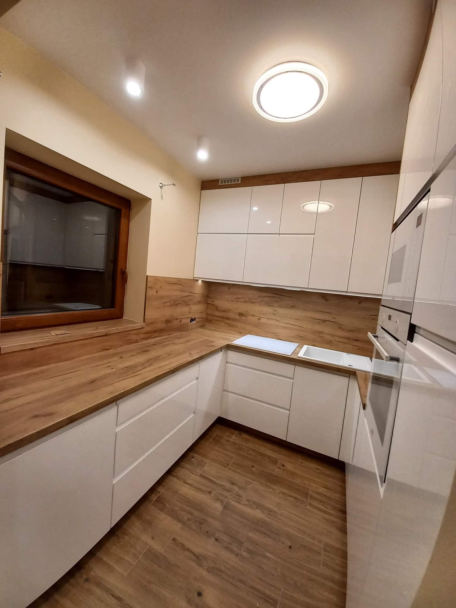 Nowoczesna kuchnia z białymi, lakierowanymi frontami, drewnianym blatem i brązowymi panelami ściennymi imitującymi drewno, oświetlona okrągłą lampą sufitową.