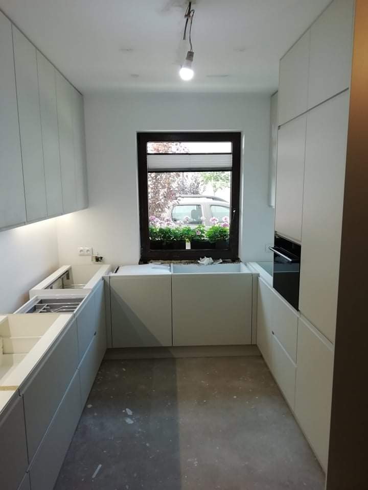 Wnętrze kuchni w trakcie montażu: białe, minimalistyczne szafki kuchenne bez blatów, okno z widokiem na samochód i kwiaty w doniczkach, surowa podłoga, wisząca żarówka bez klosza.