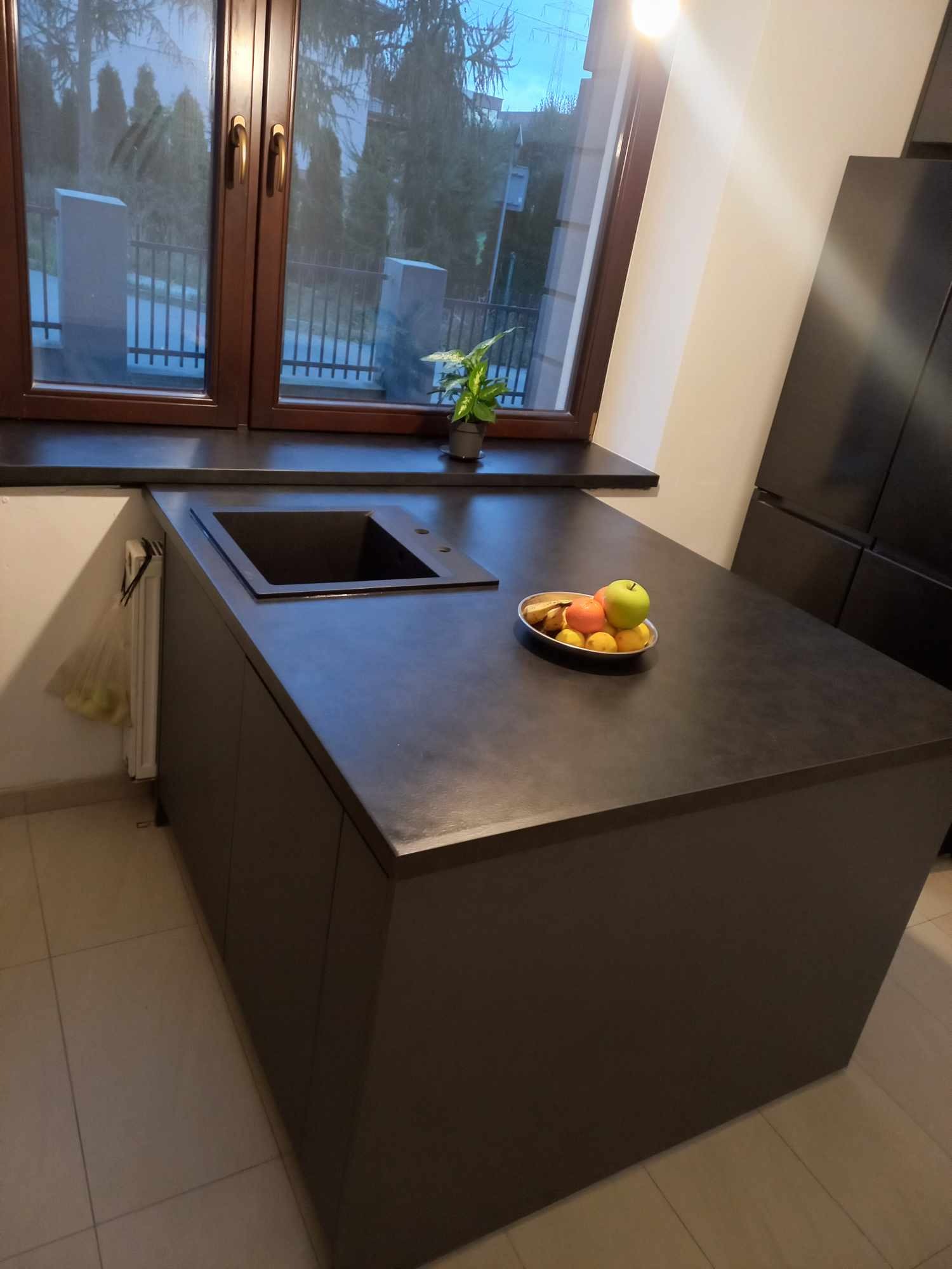 Ciemny, matowy blat kuchenny z wbudowanym zlewem i owocami, pod oknem z rośliną. Minimalistyczny design, nowoczesna kuchnia z ciemnymi meblami.