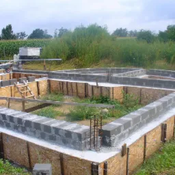 Wykop pod fundamenty domu z szalunkiem z płyt OSB, częściowo wylane ławy fundamentowe z bloczków betonowych, wystające pręty zbrojeniowe, widoczna drabina drewniana i taczka w tle.