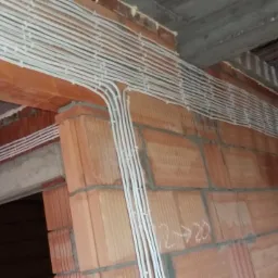 Białe przewody elektryczne ułożone równolegle na ścianie z czerwonej cegły w surowym stanie, widoczne fragmenty stropu i otwór drzwiowy.