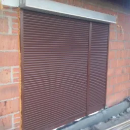 Brązowa roleta zewnętrzna zamontowana w surowym stanie na ceglanym budynku z widoczną pianką montażową i elementami budowlanymi na dole