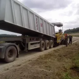 Ciężarówka z naczepą wyładowuje materiał do rozściełacza asfaltu podczas budowy drogi, w tle widoczni pracownicy i fragment ceglanego budynku.