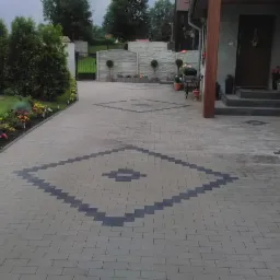 Ułożona kostka brukowa na podjeździe z geometrycznym wzorem w kształcie rombu, rabata kwiatowa wzdłuż krawędzi, dom z drewnianym filarem i schodami wejściowymi w tle.