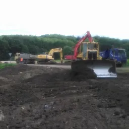 Żółty spychacz z uniesioną łyżką, koparka i dwie ciężarówki na placu budowy z usypaną ziemią, w tle zielony las i pochmurne niebo.