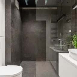 Nowoczesna łazienka z ciemnymi płytkami imitującymi kamień, kabiną prysznicową typu walk-in z czarnym sufitem i oświetleniem punktowym, białą szafką z umywalką nablatową oraz wiszącą toaletą.
