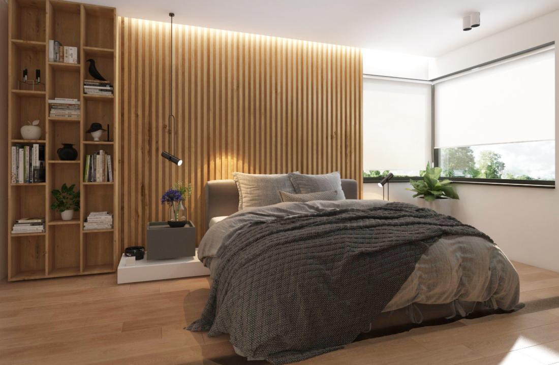 Nowoczesna sypialnia z drewnianą ścianą z lameli podświetloną od góry, drewniany regał z książkami i dekoracjami, łóżko z szarą narzutą, stolik nocny z kwiatami i lampką, rolety w oknach.