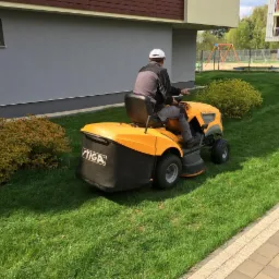 Żółta kosiarka traktorek Stiga w trakcie koszenia trawy na zielonym trawniku przed budynkiem. Widoczne równo skoszone pasy trawy i krzewy ozdobne.