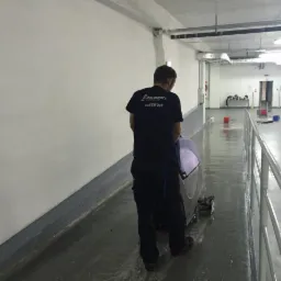 Pracownik w ciemnej koszulce z logo TopCleaner czyści posadzkę w hali przemysłowej szorowarką. Widoczne ślady wilgoci po czyszczeniu.