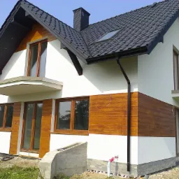 Wykończony dom jednorodzinny z drewnianą elewacją i białym tynkiem w Bielsku Podlaskim. Widoczny dach z czarnej dachówki i okna z drewnianymi ramami.