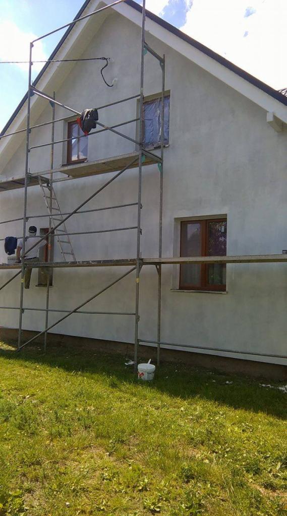 Dom w trakcie tynkowania elewacji, widoczne rusztowanie, pracownik w trakcie pracy, częściowo zakryte okna folią ochronną, wiadro z zaprawą na trawie.