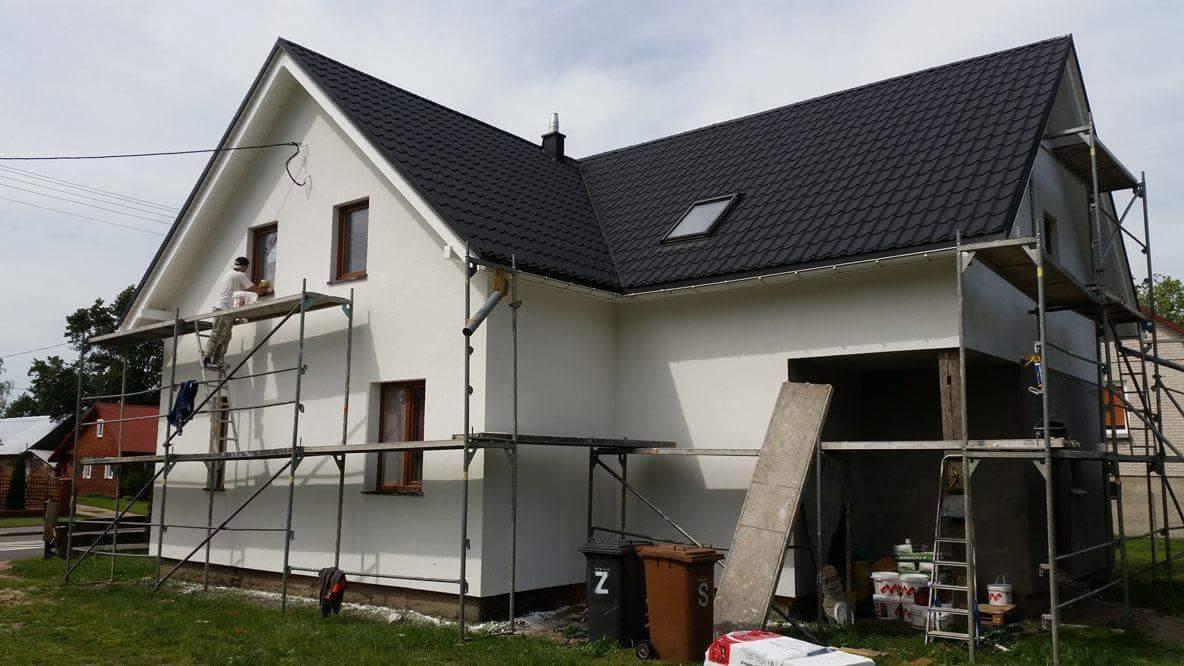 Tynkowanie elewacji domu jednorodzinnego ze rusztowania. Widoczne pędzle, farby i pojemniki. Część elewacji pomalowana na biało, część jeszcze surowa.