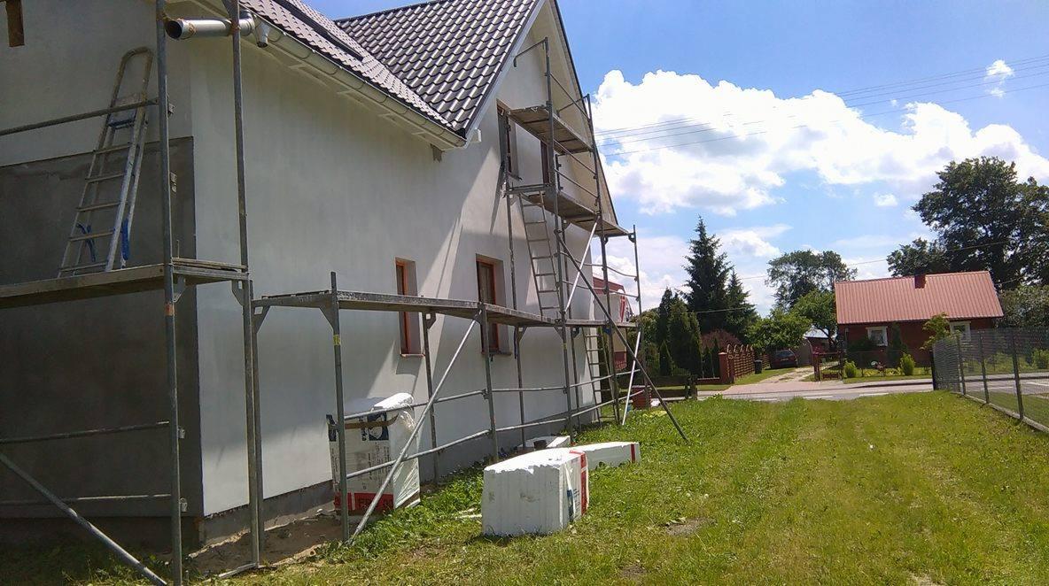 Biała elewacja domu w trakcie tynkowania, widoczne rusztowania, materiały budowlane i fragment ogrodu w słoneczny dzień. Częściowo otynkowana ściana.