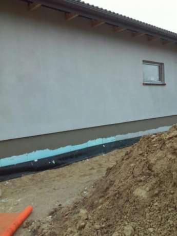 Ocieplona ściana budynku z fragmentem okna i widocznym gruntem. Prace izolacyjne w toku, widoczna warstwa izolacji termicznej i folii.