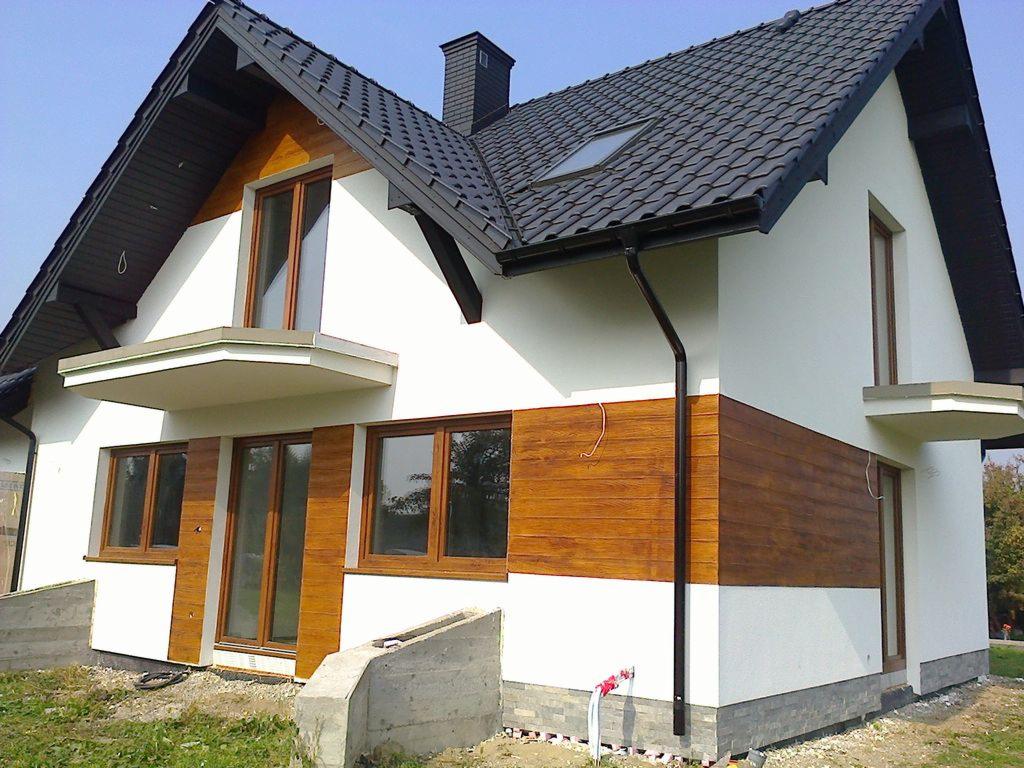 Wykończony dom jednorodzinny z drewnianą elewacją i białym tynkiem w Bielsku Podlaskim. Widoczny dach z czarnej dachówki i okna z drewnianymi ramami.