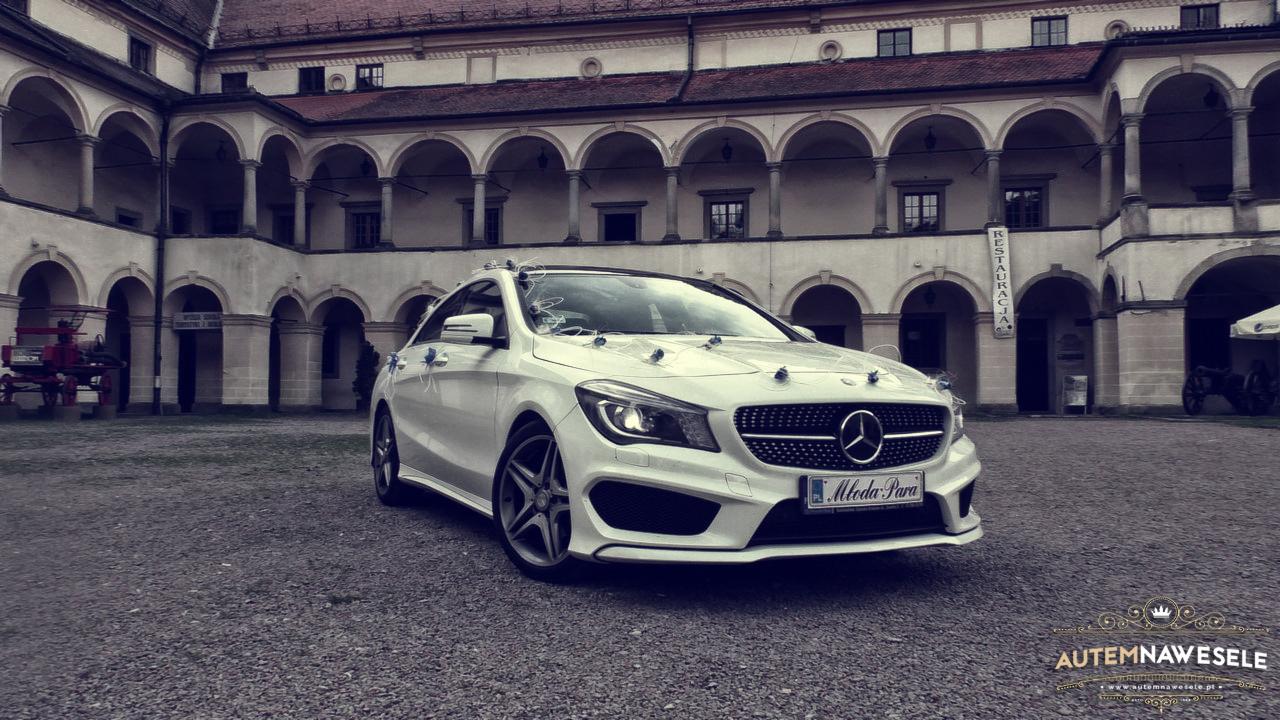Elegancki, biały Mercedes CLA z dekoracjami ślubnymi na tle arkadowego dziedzińca. Tablica rejestracyjna z napisem 'Młoda Para'.