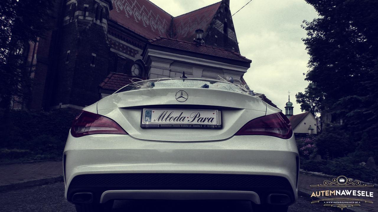 Elegancki, biały Mercedes z tablicą 'Młoda Para' na tle gotyckiego kościoła, sugerujący wynajem auta na ślub. Widok z tyłu pojazdu.