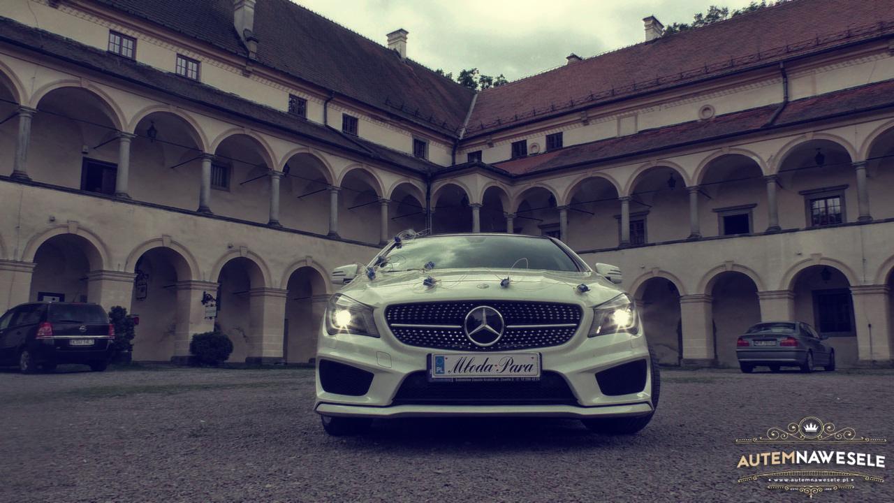 Biały Mercedes z dekoracją ślubną na masce, zaparkowany na tle dziedzińca z arkadami. Tablica rejestracyjna z napisem 'Młoda Para'.