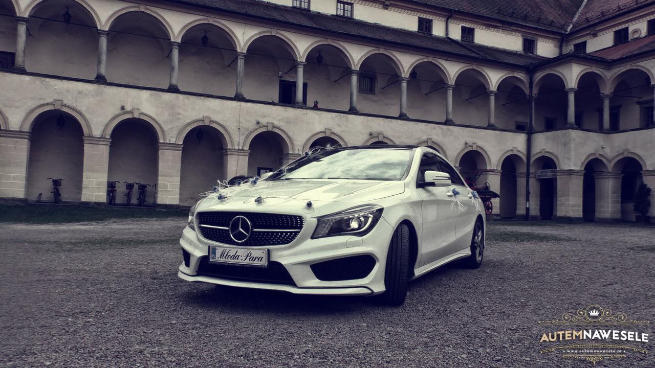 Biały Mercedes CLA udekorowany na wesele, zaparkowany na tle zabytkowego budynku z arkadami. Dekoracje kwiatowe i tablica rejestracyjna 'Młoda Para'.