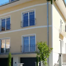 Dwukondygnacyjny budynek mieszkalny z beżową elewacją, białymi obramowaniami okien i czarnymi balkonami, widoczny z zewnątrz w słoneczny dzień.