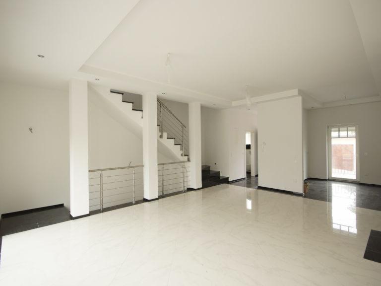 Przestronne, jasne wnętrze domu w stylu minimalistycznym z białymi ścianami, błyszczącą marmurową podłogą, schodami z metalową balustradą i dużym oknem.