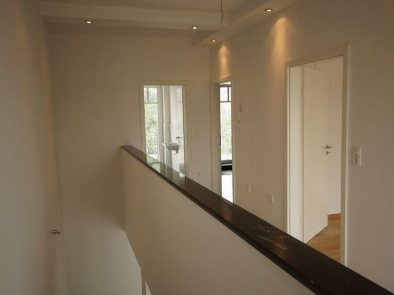 Wnętrze domu w trakcie wykańczania: białe ściany, ciemna balustrada, widoczne drzwi wewnętrzne i oświetlenie punktowe na suficie.