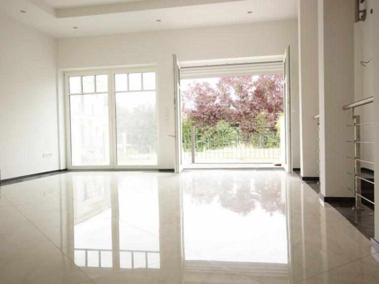 Jasne, przestronne wnętrze domu z dużymi oknami i otwartym wyjściem na ogród, polerowana podłoga z płytek odbija światło, widoczne elementy balustrady schodów.