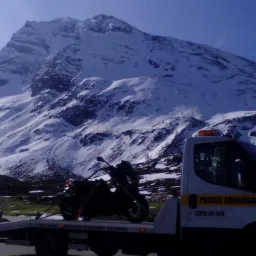 Laweta z motocyklem na tle ośnieżonych gór. Biała laweta z napisem 'Pomoc Drogowa' i numerem telefonu. Motocykl zabezpieczony pasami. Słoneczny dzień.