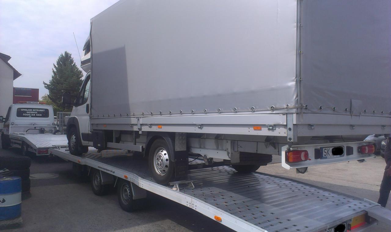 Laweta z plandeką przewożąca inny pojazd. Widoczny fragment zabudowy i drzewo w tle. Rejestracja SK.