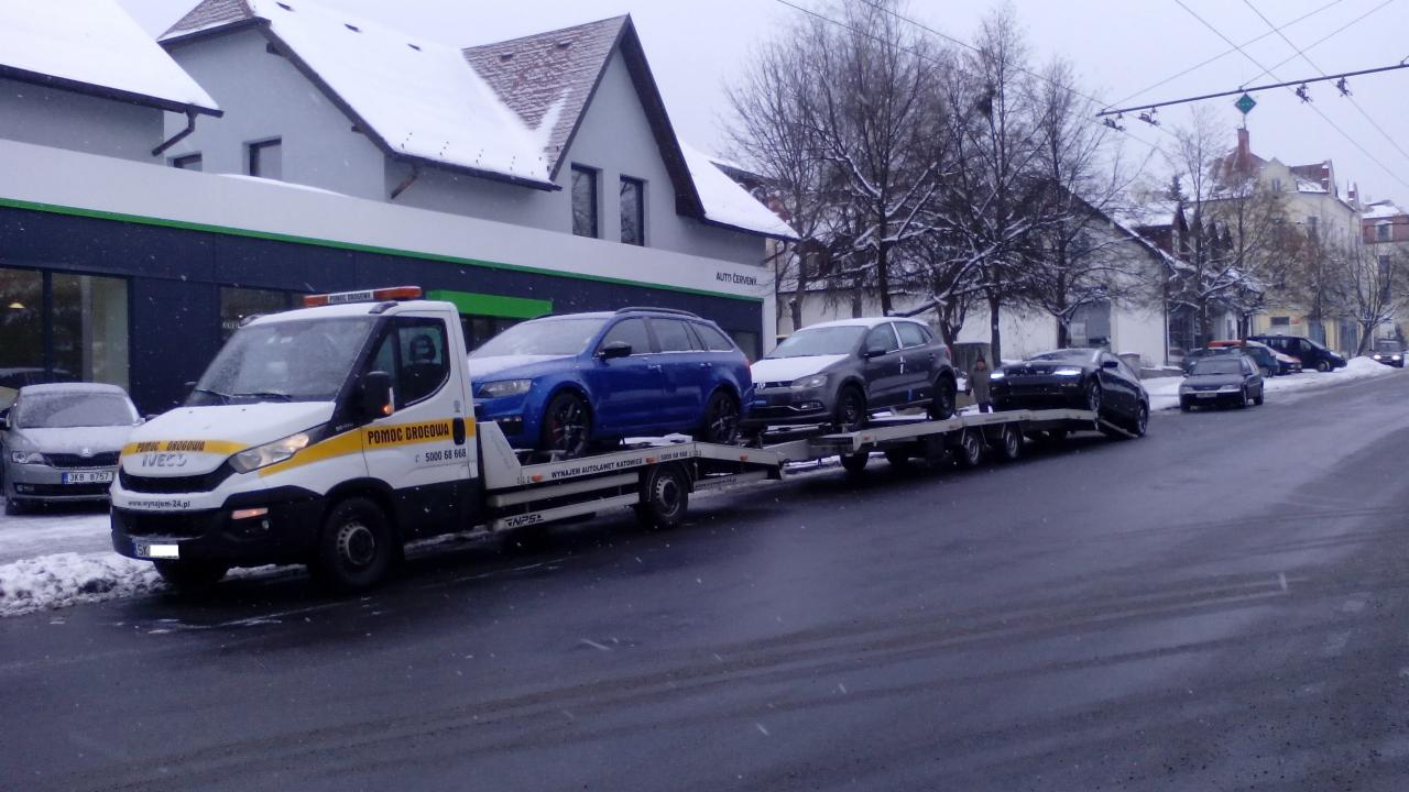 Laweta z trzema samochodami na platformie, zaparkowana przy ulicy w zimowej scenerii miejskiej. Na lawecie niebieska Skoda, szary VW i czarny samochód.