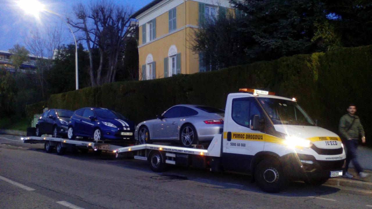 Laweta z trzema autami (Ford Fiesta, Porsche) na tle budynku z zielonymi okiennicami i wysokiego żywopłotu. Widoczny mężczyzna idący chodnikiem.